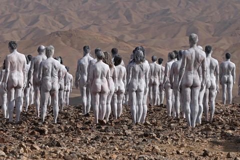 In einer braunen Wüstenlandschaft stehen nackte, weiß angemalte Menschen