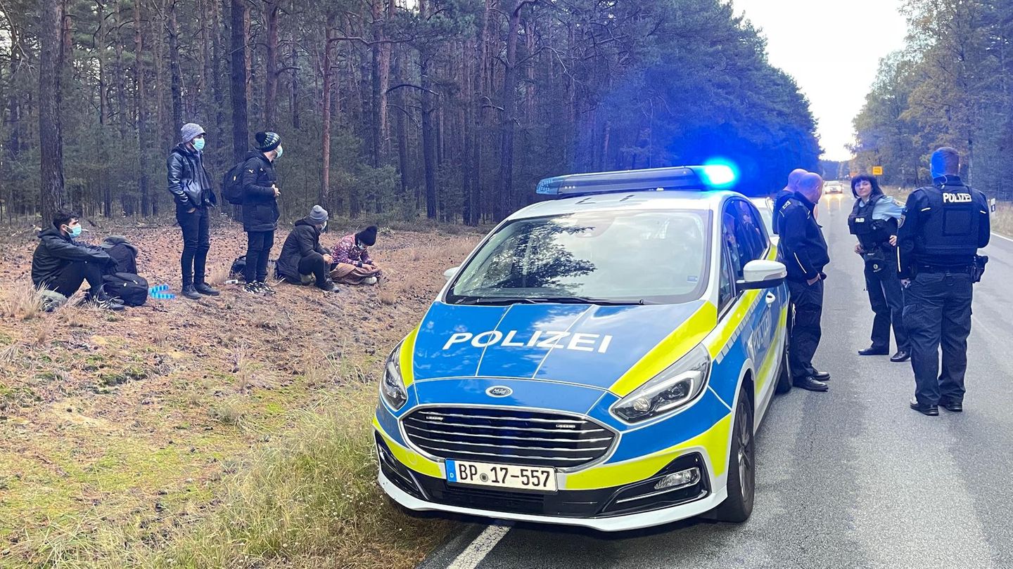 An einem Waldrand sitzen und stehen fünf Menschen, auf der Straße vor ihnen steht ein Polizeiwagen mit Blaulicht