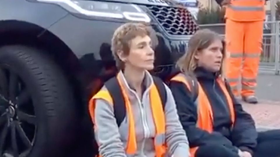 Zwei Frauen sitzen in Warnwesten auf einer Straße. Hinter ihnen steht ein schwarzer SUV