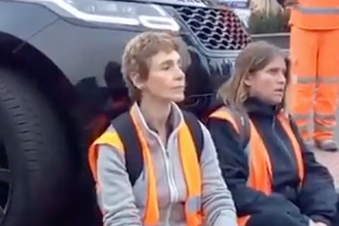 Zwei Frauen sitzen in Warnwesten auf einer Straße. Hinter ihnen steht ein schwarzer SUV