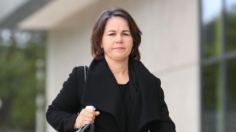 Eine weiße Frau mit schulterlangen, braunen Haaren geht mit Handtasche unter dem rechten Arm an einem hellen Gebäude vorbei