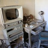 Die Inneneinrichtung einer verlassenen Wohnung. Neben einem Schreibtisch samt Stuhl steht ein altes Fernsehgerät. Eine Tasse und ein kleines Kännchen wurden dort einfach zurückgelassen.