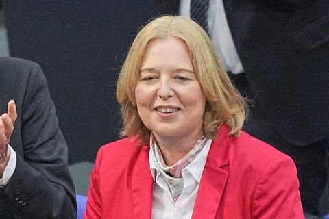 Eine weiße mittelalte Frau mit schulterlangen blonden Haaren und in rotem Blazer lächelt und legt dankend die Hände zusammen