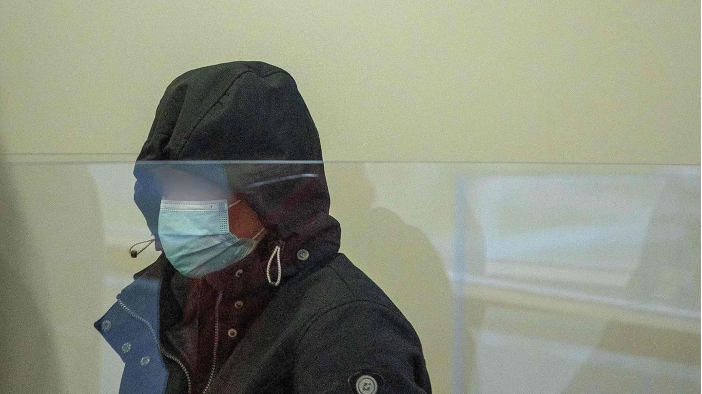 Hinter einer Plexiglasscheibe sitzt eine Frau, die sich in einer dunkelblauen Kapuze und hinter einer OP-Maske verbirgt