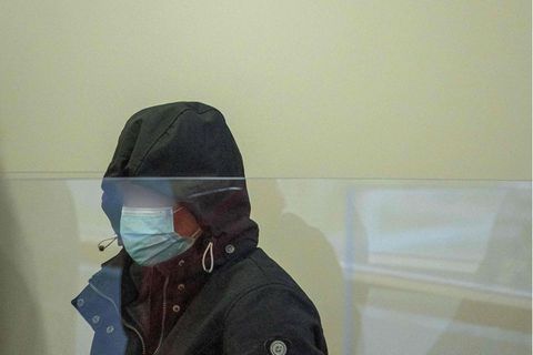 Hinter einer Plexiglasscheibe sitzt eine Frau, die sich in einer dunkelblauen Kapuze und hinter einer OP-Maske verbirgt