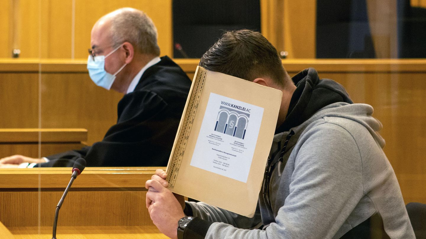 Hinter einer beigen Mappe verbirgt ein junger weißer Mann sein Gesicht. Er hat einen gegelten braunen Seitenscheitel