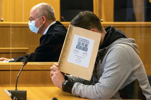 Hinter einer beigen Mappe verbirgt ein junger weißer Mann sein Gesicht. Er hat einen gegelten braunen Seitenscheitel