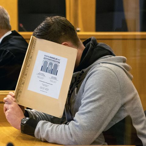 Hinter einer beigen Mappe verbirgt ein junger weißer Mann sein Gesicht. Er hat einen gegelten braunen Seitenscheitel