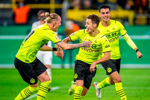 Drei Fußballer in gelben Trikots und schwarzen kurzen Hosen laufen jubelnd über einen Fußballplatz