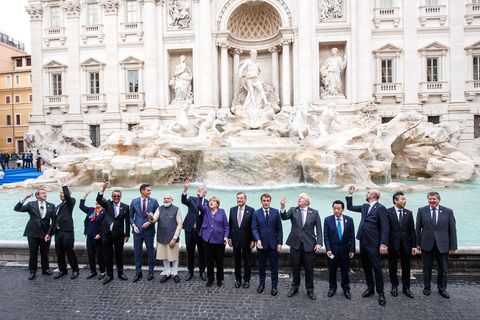Eine Gruppe Männer n Anzügen, ein Mann in indischer Kleidung sowie Angela Merkel in einem lila Blazer stehen vor einem Brunnen