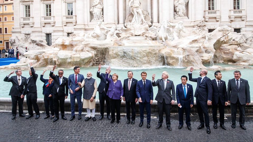 Eine Gruppe Männer n Anzügen, ein Mann in indischer Kleidung sowie Angela Merkel in einem lila Blazer stehen vor einem Brunnen