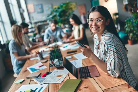 Frau sitzt mit anderen Personen gemeinsam am Tisch. Alle arbeiten an Laptops oder auf Papier.