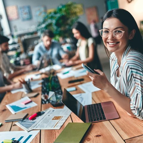 Frau sitzt mit anderen Personen gemeinsam am Tisch. Alle arbeiten an Laptops oder auf Papier.