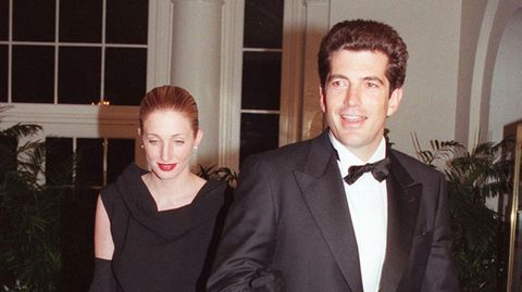 Ein weißer Mann in Anzug und Fliege und eine weiße, blonde Frau im schwarzen Abendkleid gehen untergehakt
