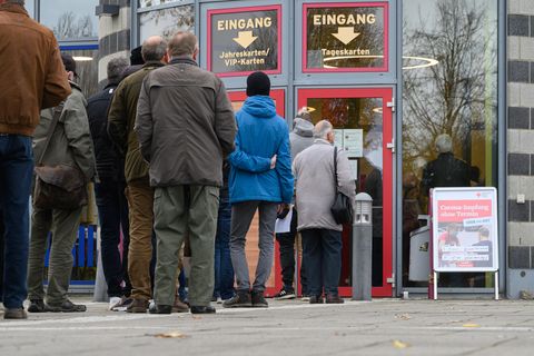 Vor Glastüren mit roten Rahmen stehen Menschen in warmer Kleidung Schlange