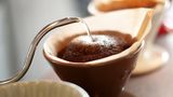 Der Aufguss  Schritt 5: Niemals den Kaffee mit kochendem Wasser übergießen!  Der von Hand aufgebrühte Kaffee schmeckt aromatischer und runder, wenn das Wasser nicht mehr kocht, wenn es mit ihm in Berührung kommt. Bei zu großer Hitze verflüchtigen sich die Aromastoffe und die bitteren und sauren Komponenten treten in den Vordergrund. Ideal ist eine Temperatur von 86 bis 89 Grad Celsius. Das Kaffeemehl nun mit einem kleinen Schwall Wasser anfeuchten und es 20 Sekunden quellen lassen. Auf diese Weise werden die gerösteten Öle, Fette und Bitterstoffe gelöst. Nun das Wasser in kleinen Mengen alle zehn Sekunden schwallweise nachgießen. Der Vorgang sollte vier bis sechs Minuten dauern. Unter vier Minuten entfalten sich die Aromen nicht, über sechs Minuten verflüchtigen sie sich bereits. Das manuelle Aufbrühen ist auch heute noch die Methode, mit der Kaffeetester die Qualität der Ware beurteilen.