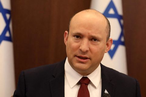 Ein weißer Mann mit Halbglatze und rundlichem Gesicht sitzt in Anzug und Krawatte vor zwei israelischen Flaggen