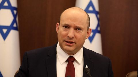 Ein weißer Mann mit Halbglatze und rundlichem Gesicht sitzt in Anzug und Krawatte vor zwei israelischen Flaggen