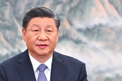 Ein chinesisch aussehender Mann mit angegrauten Schläfen und Seitenscheitel sitzt in Anzug und Krawatte vor einem Bergpanorama