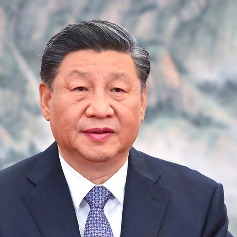 Ein chinesisch aussehender Mann mit angegrauten Schläfen und Seitenscheitel sitzt in Anzug und Krawatte vor einem Bergpanorama
