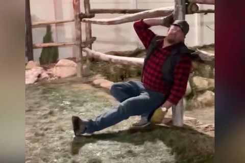 Ein Holzfäller lehnt rückwärts in der Hocke an einem Holzstamm.