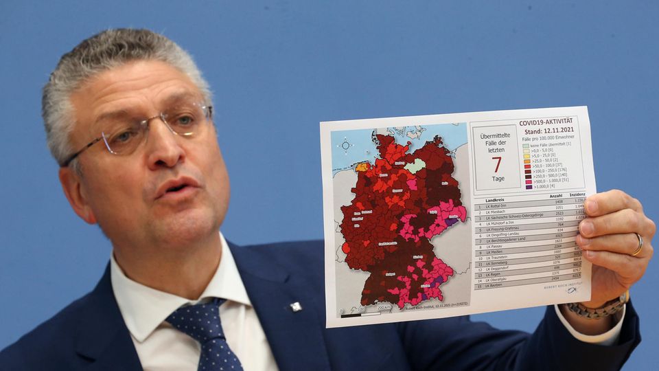 Eoin weißer Mann mit weißgrauen Locken hält mit links eine Deutschlandkarte mit unterschiedlich rot gefärbten Landkreisen hoch