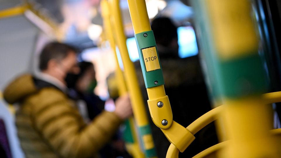 Ein weißer Mann mit schwarzer FFP2-Maske und brauner Steppjacke hält sich in einem Bus an gelbem Gestänge fest