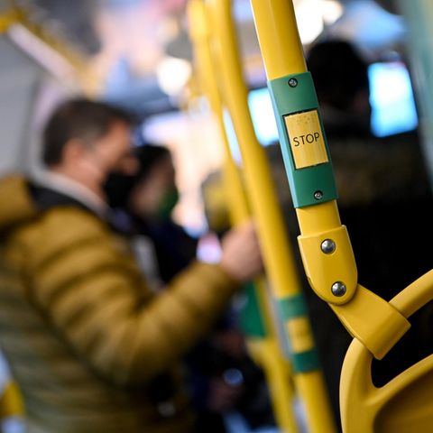 Ein weißer Mann mit schwarzer FFP2-Maske und brauner Steppjacke hält sich in einem Bus an gelbem Gestänge fest