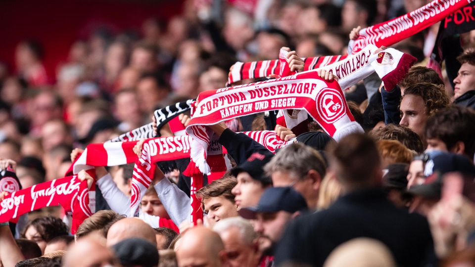 Aus einer Menschenmasse in einem Stadion recken Arme rot-weiße Fanschals von Fortuna Düsseldorf hoch