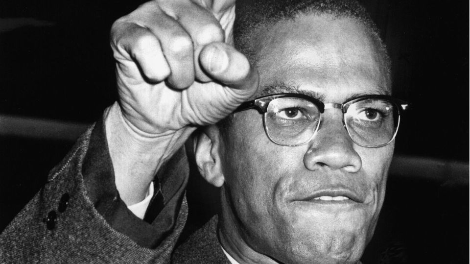 Malcolm X blickt entschlossen und recht eine Hand in die Höhe