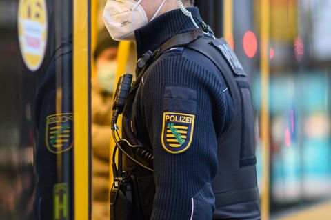 Ein weißer Mann in dunkelblauer Polizeiuniform und Wappen von Sachsen auf dem Arm steht an der Tür einer gelben Straßenbahn