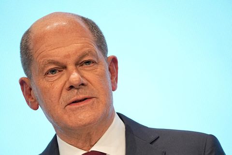Ein weißer Mann mit Halbglatze steht in Anzug und Krawatte vor einem hellblauen Hintergrund und spricht