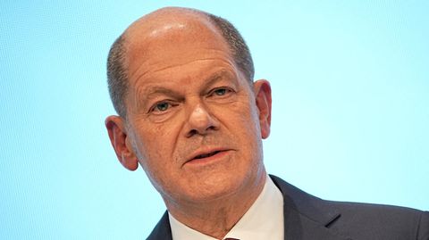 Ein weißer Mann mit Halbglatze steht in Anzug und Krawatte vor einem hellblauen Hintergrund und spricht