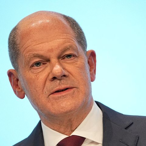 Ein weißer Mann mit Halbglatze steht in Anzug und Krawatte vor einem hellblauen Hintergrund und spricht