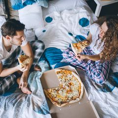 Ein Paar sitzt in Pyjamas im Bett und isst Pizza