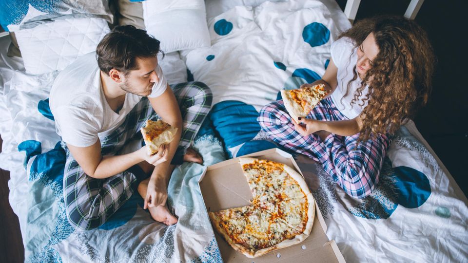 Ein Paar sitzt in Pyjamas im Bett und isst Pizza