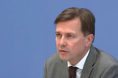 Ein mittelalter weißer Mann mit braunem Seitenscheitel sitzt in graugrünem Jackett an einem hellen Holzpodium