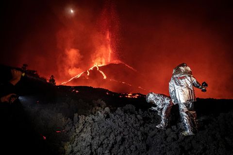 Ein rot glühender Vulkan im Hintergrund. Im Vordergrund Menschen in silbernen Schutzanzügen
