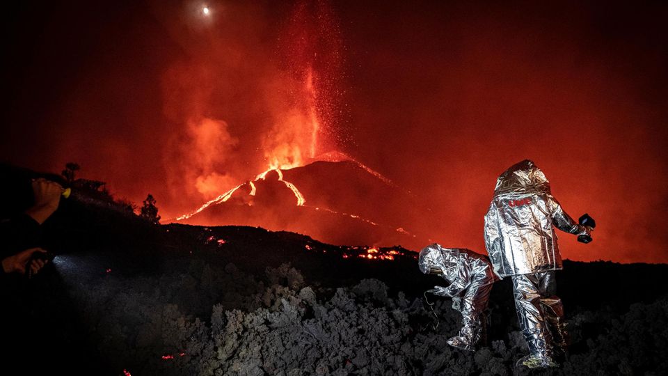 Ein rot glühender Vulkan im Hintergrund. Im Vordergrund Menschen in silbernen Schutzanzügen