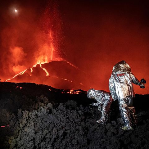 Ein rot glühender Vulkan im Hintergrund. Im Vordergrund Menschen in silbernen Schutzanzügen