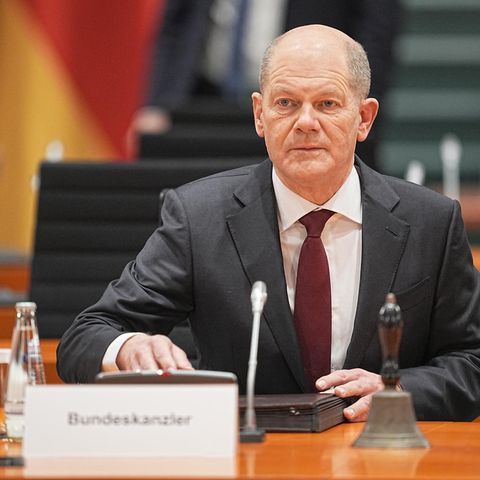 Ein weißer älterer Mann im Anzug und mit Halbglatze sitzt in Anzug und Krawatte hinter einem "Bundeskanzler"-Schild am Tisch