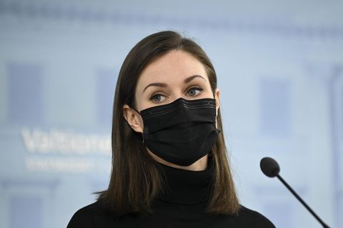 Eine junge weiße Frau mit langem braunem Haar steht mit schwarzer FFP2-Maske und Rollkragen-Pullover vor einem Mikrofon
