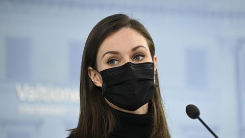 Eine junge weiße Frau mit langem braunem Haar steht mit schwarzer FFP2-Maske und Rollkragen-Pullover vor einem Mikrofon