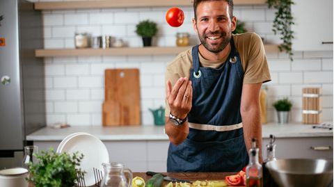 Ein Mann in Kochschürze lächelt in die Kamera während er eine Tomate hochwirft