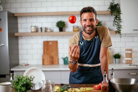 Ein Mann in Kochschürze lächelt in die Kamera während er eine Tomate hochwirft