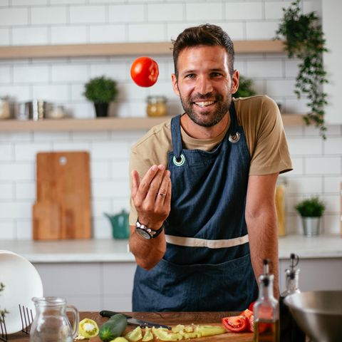 Ein Mann in Kochschürze lächelt in die Kamera während er eine Tomate hochwirft