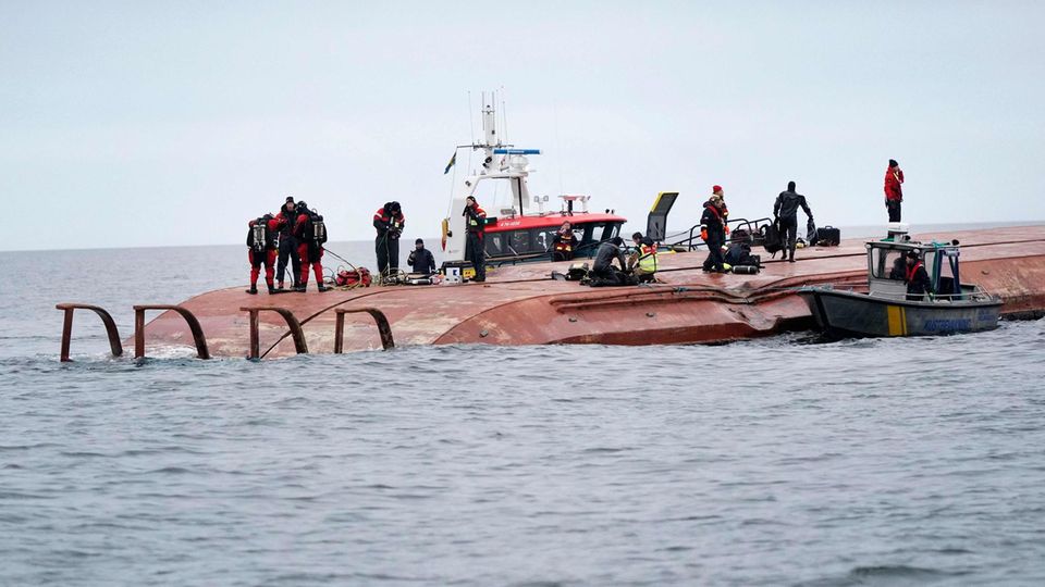 Karin Hoej Schiff gekentert Auf einem rostbraunen Schiffsrumpf, der aus dem Wasser ragt, laufen Taucher und Rettungskräfte, während