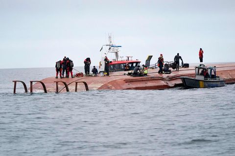 Auf einem rostbraunen Schiffsrumpf, der aus dem Wasser ragt, laufen Taucher und Rettungskräfte, während
