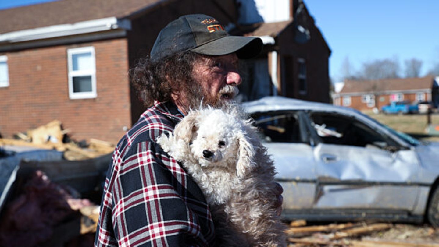 Mann mit Hund auf dem Arm steht vor zerstörtem Auto