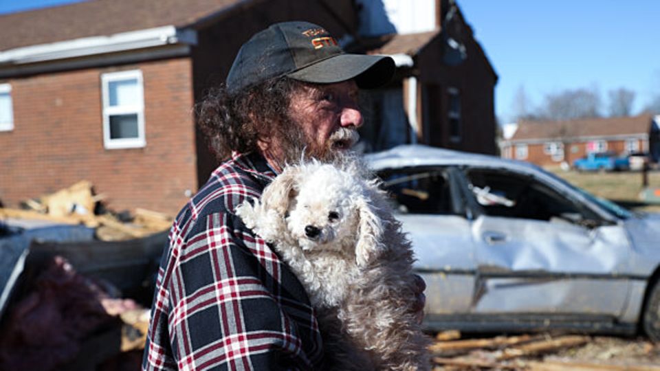 Mann mit Hund auf dem Arm steht vor zerstörtem Auto